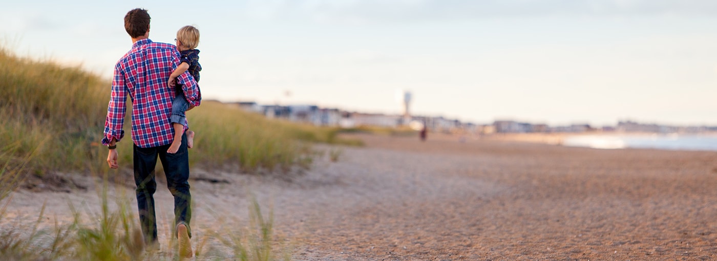 Young dad carries his son across a beach.